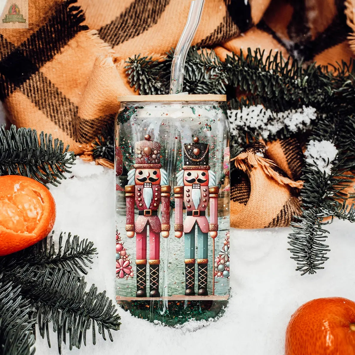 The Nutcracker Glam Soldier tumbler stands in snow with pine branches, a plaid blanket, and tangerines. Featuring two painted nutcrackers and a clear straw, it's festive holiday drinkware perfect for the season.