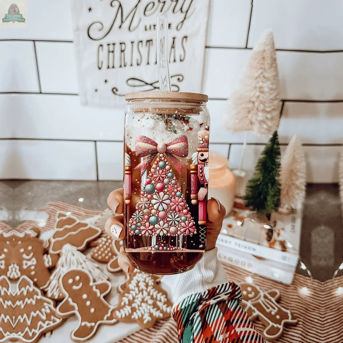A hand holds the Nutcracker Glam Soldier holiday drinkware jar with a colorful Christmas tree design; gingerbread cookies, faux trees, and a Merry Christmas sign create a cozy, festive background.