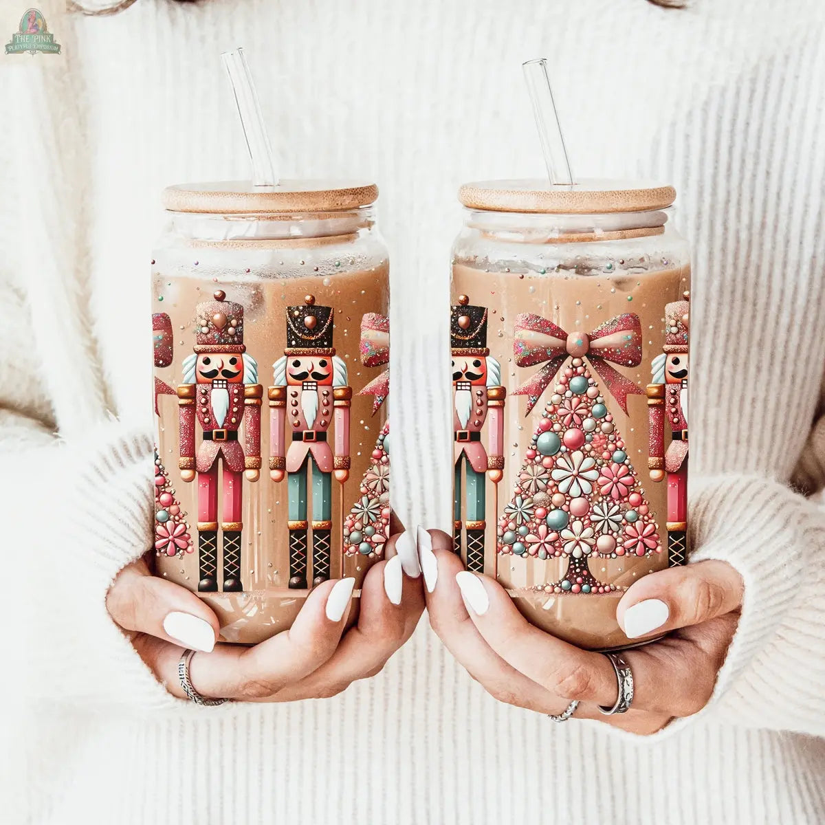 Two hands hold festive iced coffee jars featuring Nutcracker Glam Soldier holiday designs and a Christmas tree. The person wears a cozy white sweater and has neatly manicured white nails.