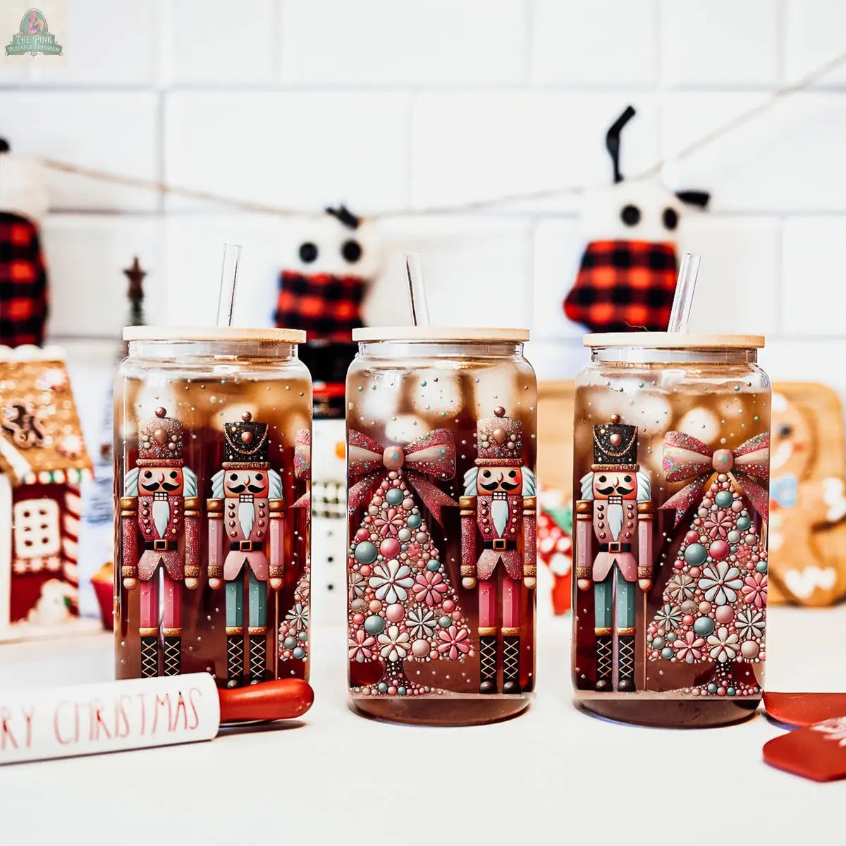 Three glass jars, including the Nutcracker Glam Soldier tumbler, are filled with iced drinks and topped with wooden lids and straws. Holiday decorations and a gingerbread house add festive cheer to this holiday drinkware collection.