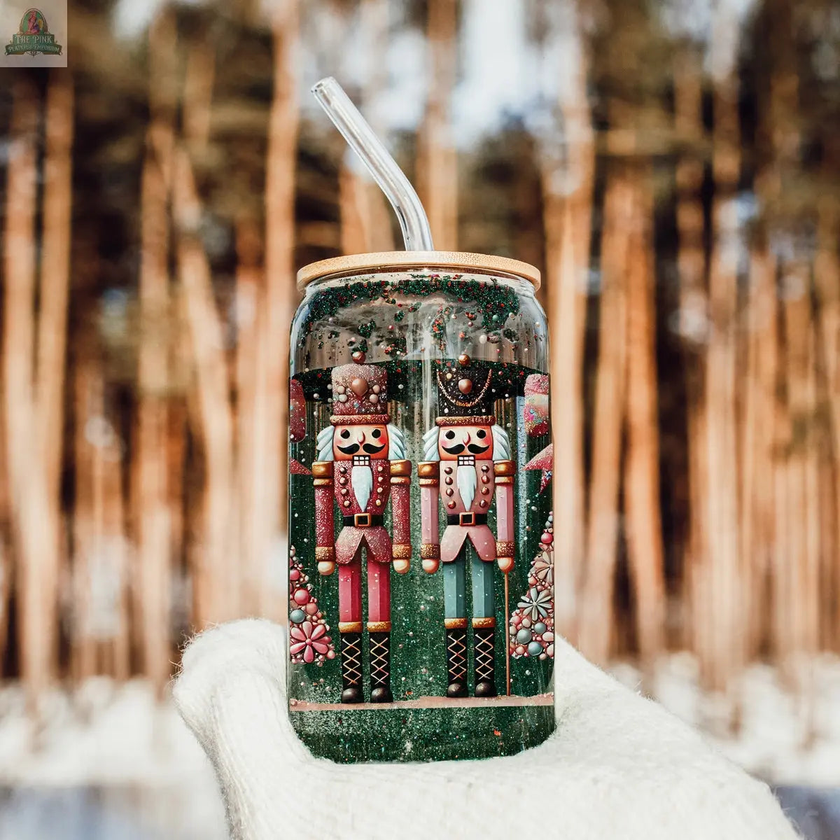 The Nutcracker Glam Soldier glass cup with a clear straw showcases two festive nutcracker soldier designs—perfect for your holiday drinkware collection. Snow and blurred tall trees set a charming outdoor background.