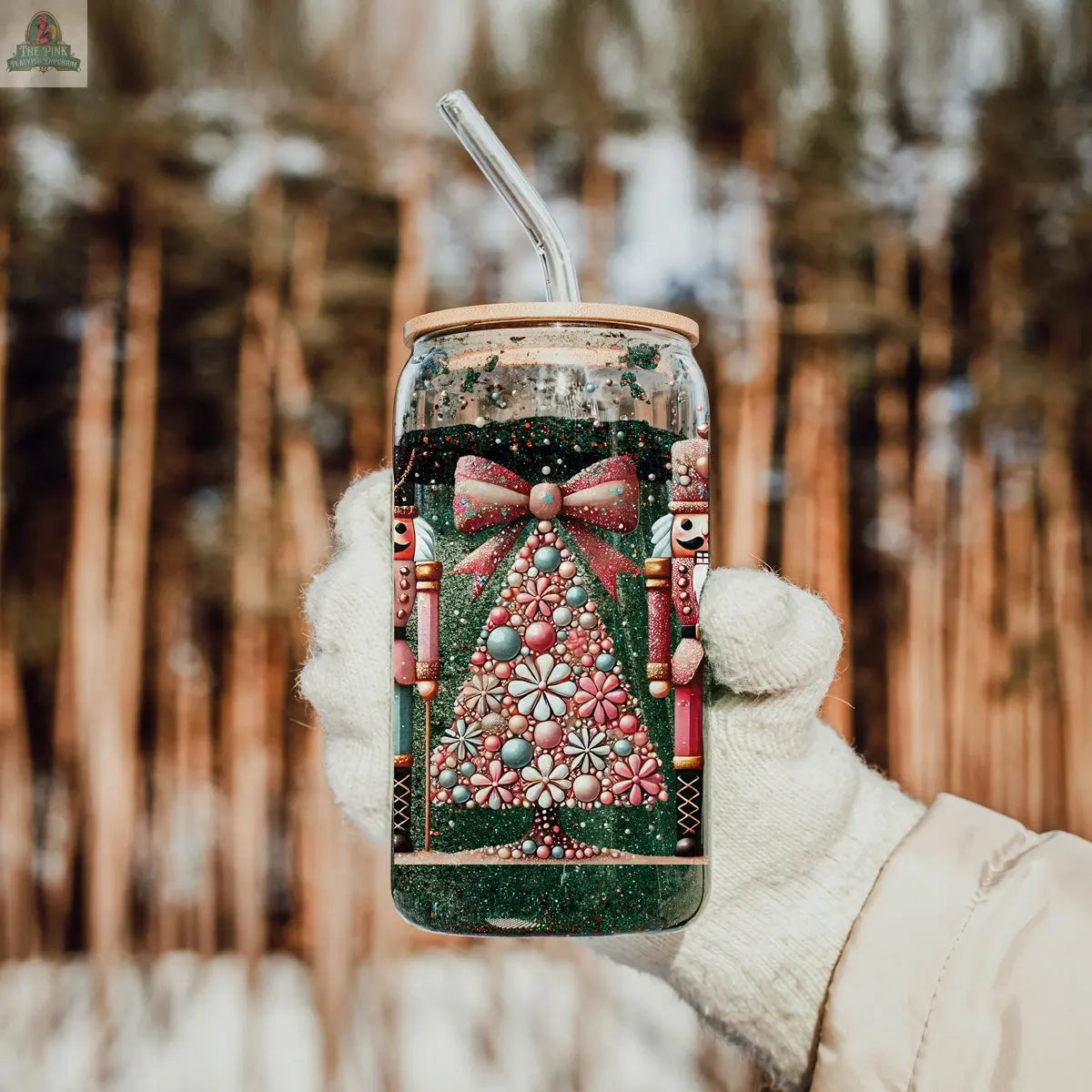 A gloved hand holds the Nutcracker Glam Soldier Christmas glass tumbler with a straw, featuring tree, bow, and nutcracker designs. Snow-dusted trees and a blurred winter forest add charm to this festive holiday drinkware scene.