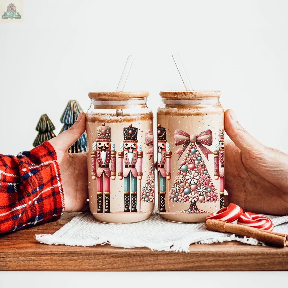 Two hands hold jars of a creamy drink with clear straws. The Nutcracker Glam Soldier tumbler features festive holiday designs, while candy canes and miniature Christmas trees decorate the background for extra charm.