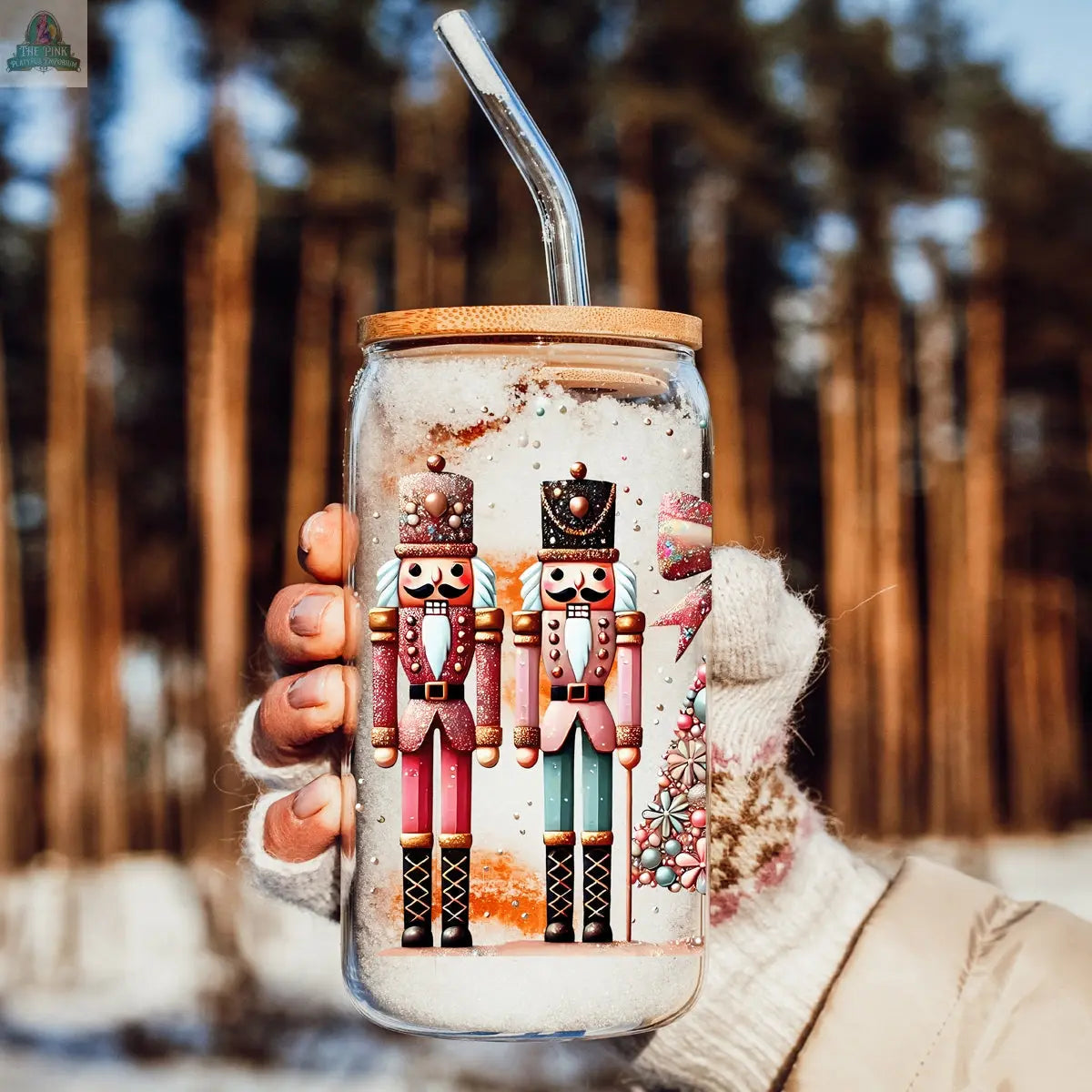 A gloved hand holds the Nutcracker Glam Soldier tumbler with a bamboo lid and straw, featuring colorful designs and snowflake patterns against a snowy forest background—the perfect holiday drinkware.