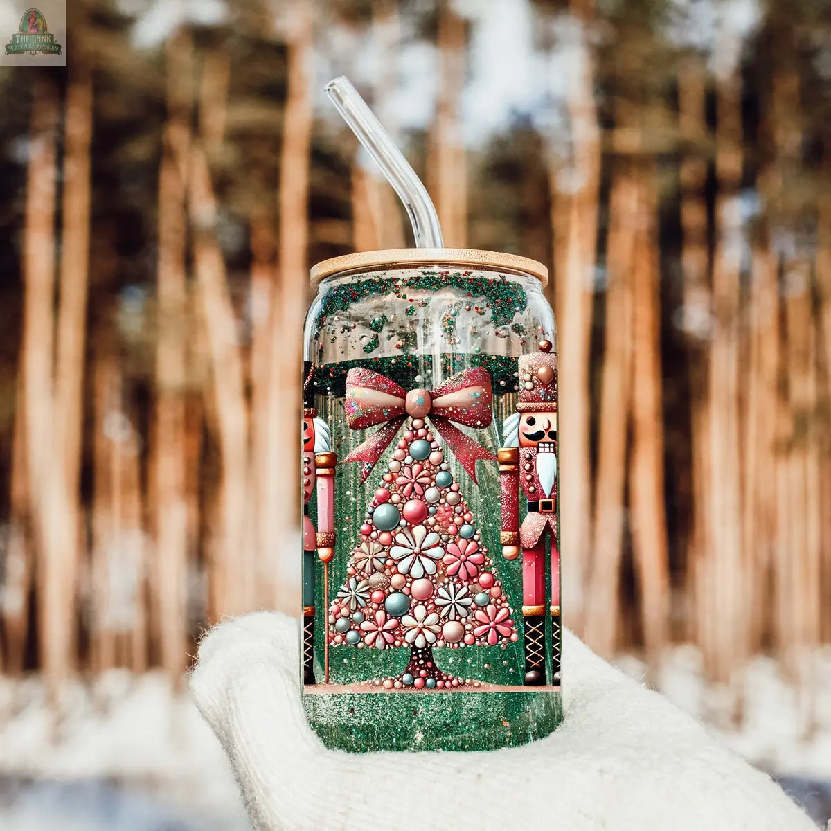 A gloved hand holds the Nutcracker Glam Soldier tumbler with a bamboo lid and straw, featuring jeweled Christmas tree and nutcracker designs. This festive holiday drinkware stands out against a backdrop of tall, blurred forest trees.