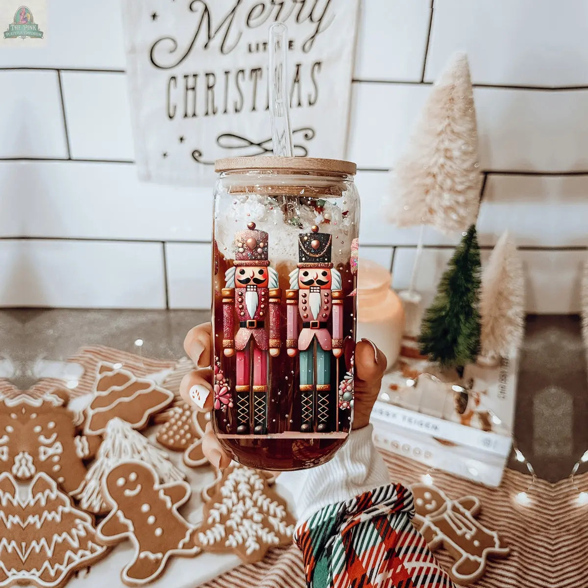A hand holds a festive holiday drink in a Nutcracker Glam Soldier tumbler, surrounded by gingerbread cookies, tabletop Christmas trees, and a "Merry Christmas" sign for a cozy holiday vibe.