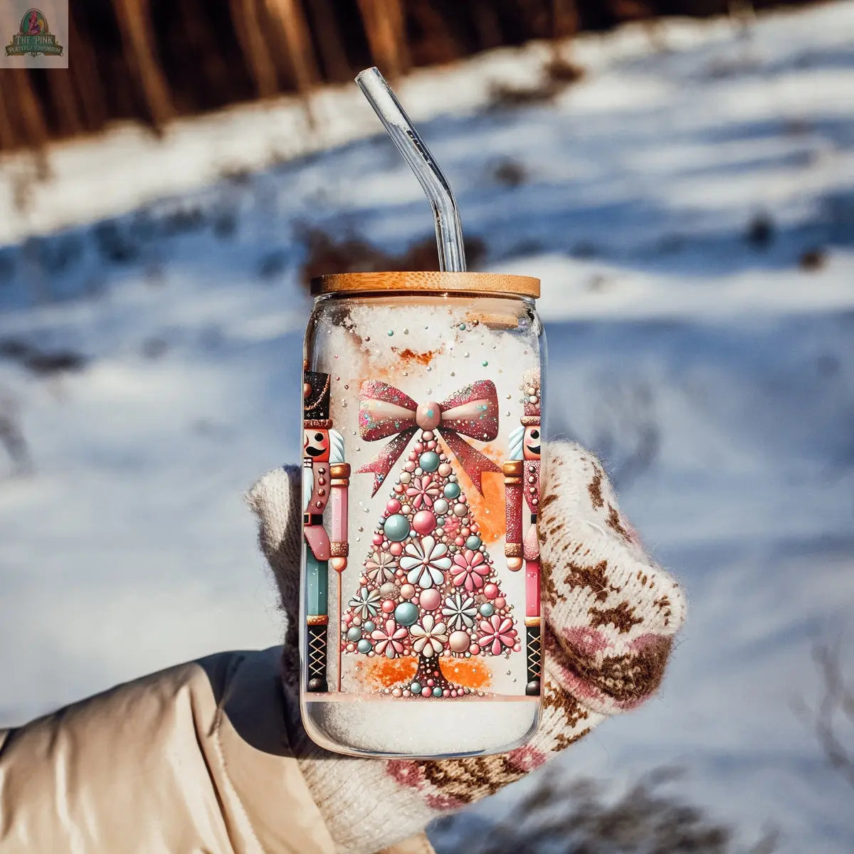 A mittened hand holds the Nutcracker Glam Soldier glass tumbler with bamboo lid and straw, featuring a jeweled Christmas tree, pink bow, and nutcrackers. Snow and trees in the background add a festive touch.