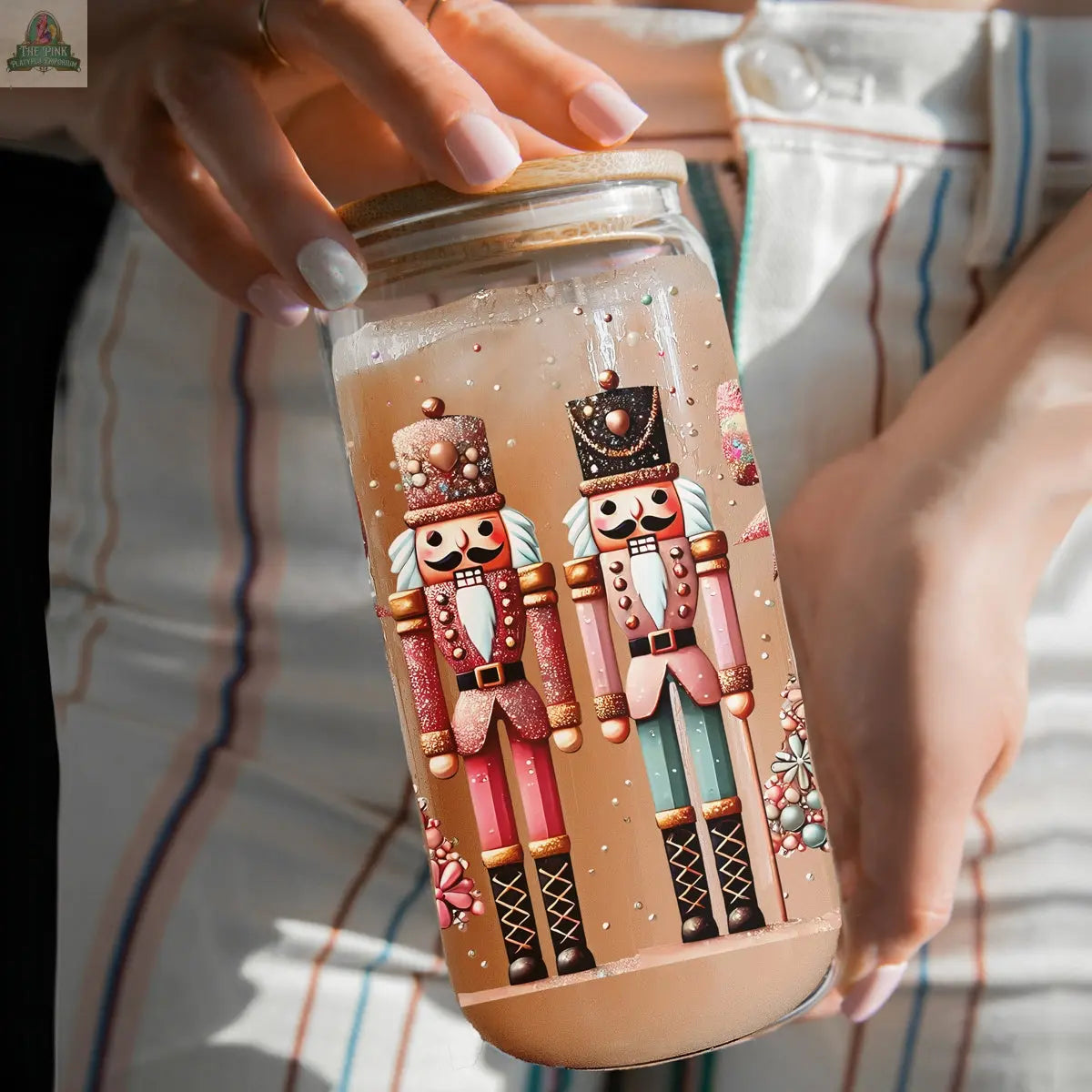 A person holds a Nutcracker Glam Soldier glass tumbler filled with a light brown drink; the tumbler features two colorful nutcracker figures in striped white pants and festive ornaments, adding charm to holiday drinkware.