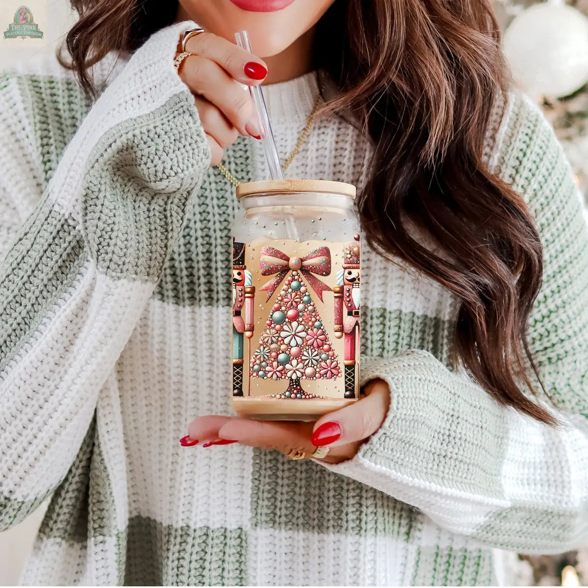 A woman with long hair and red nails sips from a Nutcracker Glam Soldier Christmas glass tumbler, featuring a festive tree, nutcracker design, and bow, while wearing a green and white striped sweater.