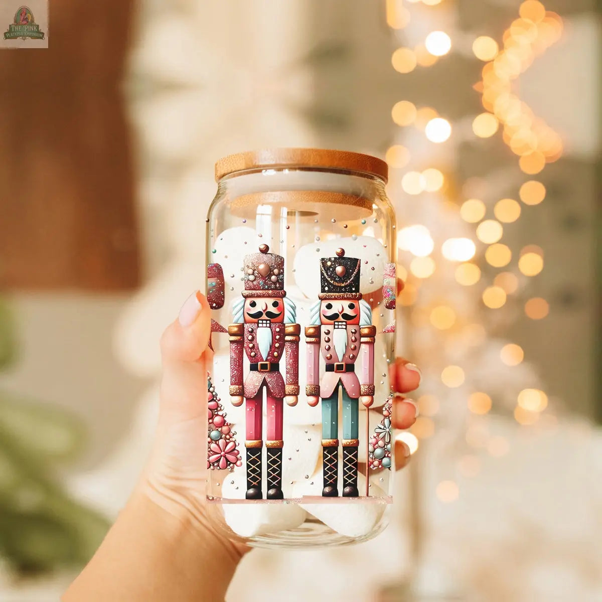 A hand holds the Nutcracker Glam Soldier Christmas glass tumbler with a bamboo lid, featuring colorful Nutcracker Soldier art and festive ornaments. Warm, blurred holiday lights twinkle in the background—perfect for your holiday drinkware.