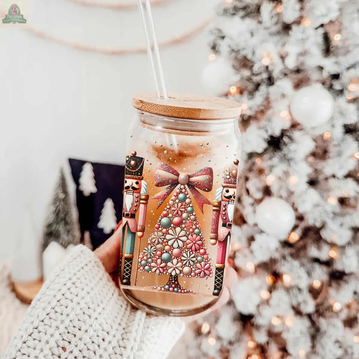 A hand in a cozy white sweater holds the Nutcracker Glam Soldier tumbler with a bamboo lid and straw, featuring nutcrackers and a Christmas tree. A blurred, lit Christmas tree with snowy decor is in the background.