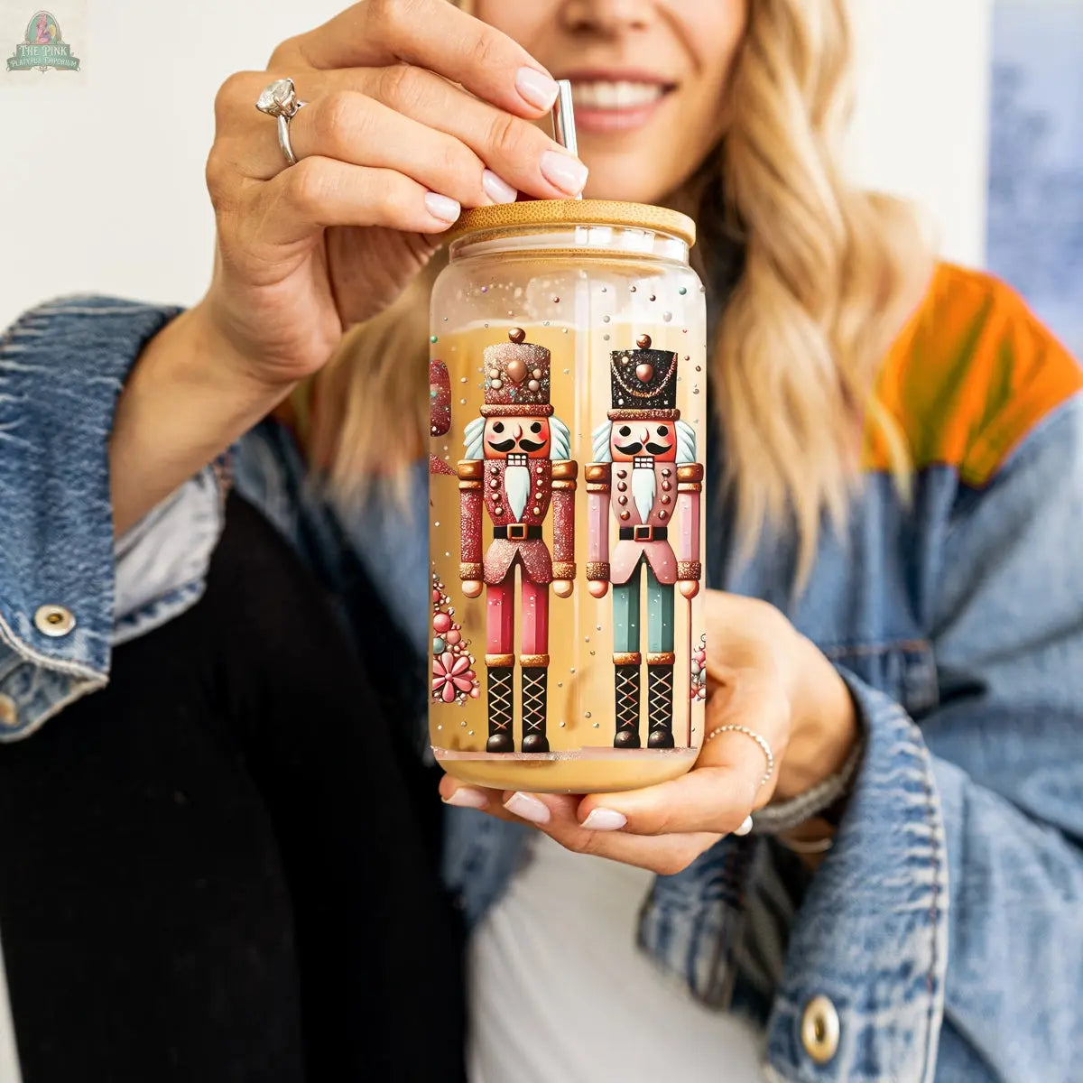 A person wearing a denim jacket and ring holds a Nutcracker Glam Soldier Christmas glass tumbler, decorated with two nutcrackers and filled with a light brown drink, sipping from a straw in the festive holiday drinkware.