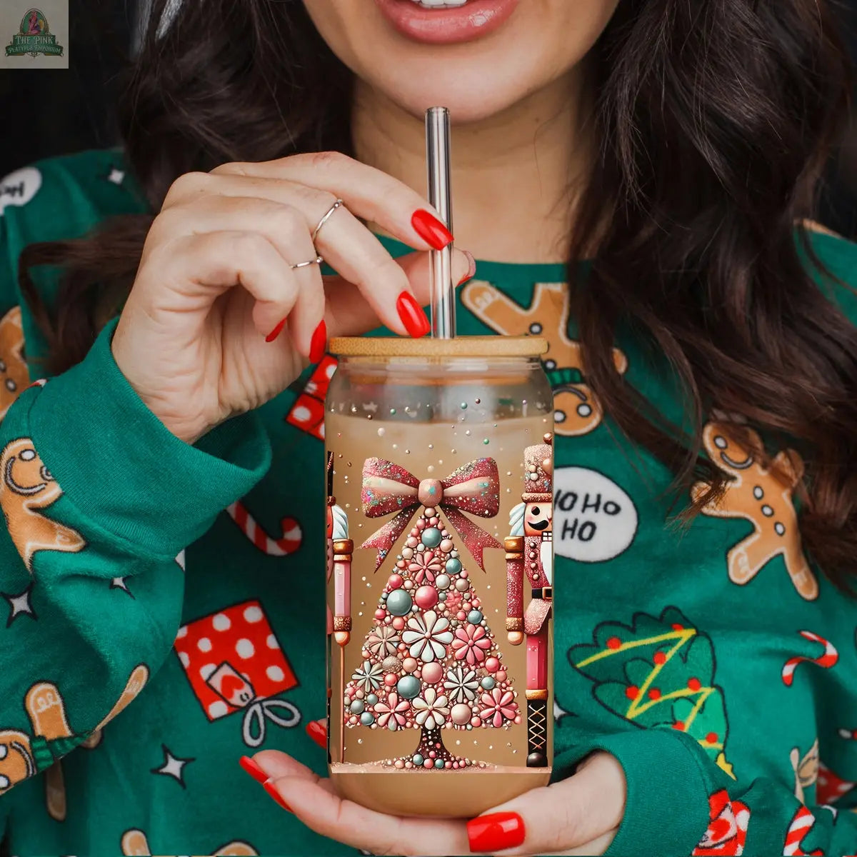 A woman in a green holiday sweater with gingerbread men and gifts sips iced coffee from the Nutcracker Glam Soldier Christmas glass tumbler, topped with a Christmas tree, candy canes, and a pink bow, using a metal straw.