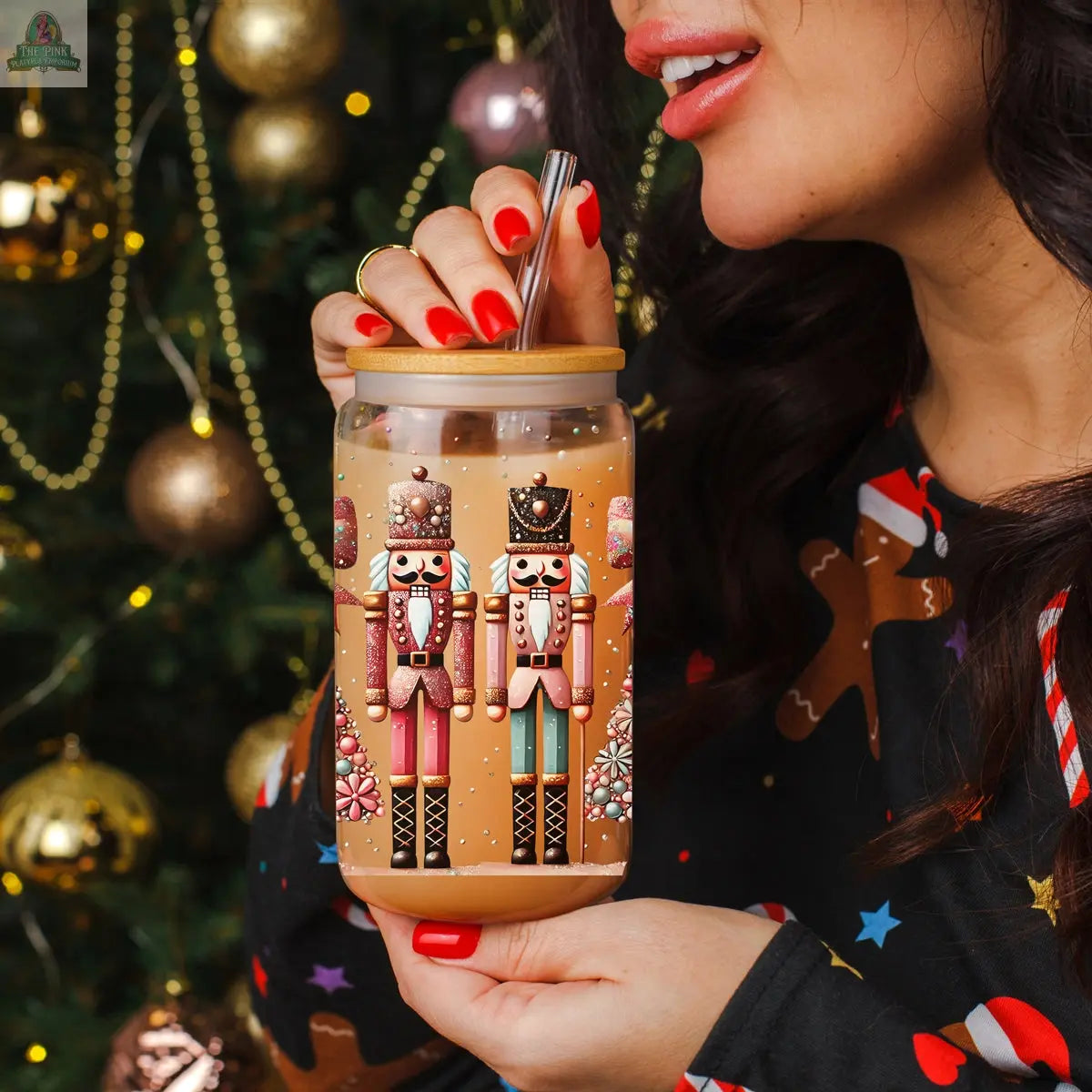 A woman with red nail polish sips from a Nutcracker Glam Soldier Christmas tumbler. A decorated tree with gold ornaments lends extra holiday charm to the festive scene.