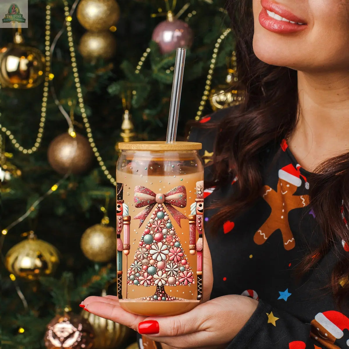 A woman in festive attire holds a Nutcracker Glam Soldier tumbler with a straw, featuring nutcrackers and a Christmas tree, standing in front of a gold-ornamented, twinkling Christmas tree.