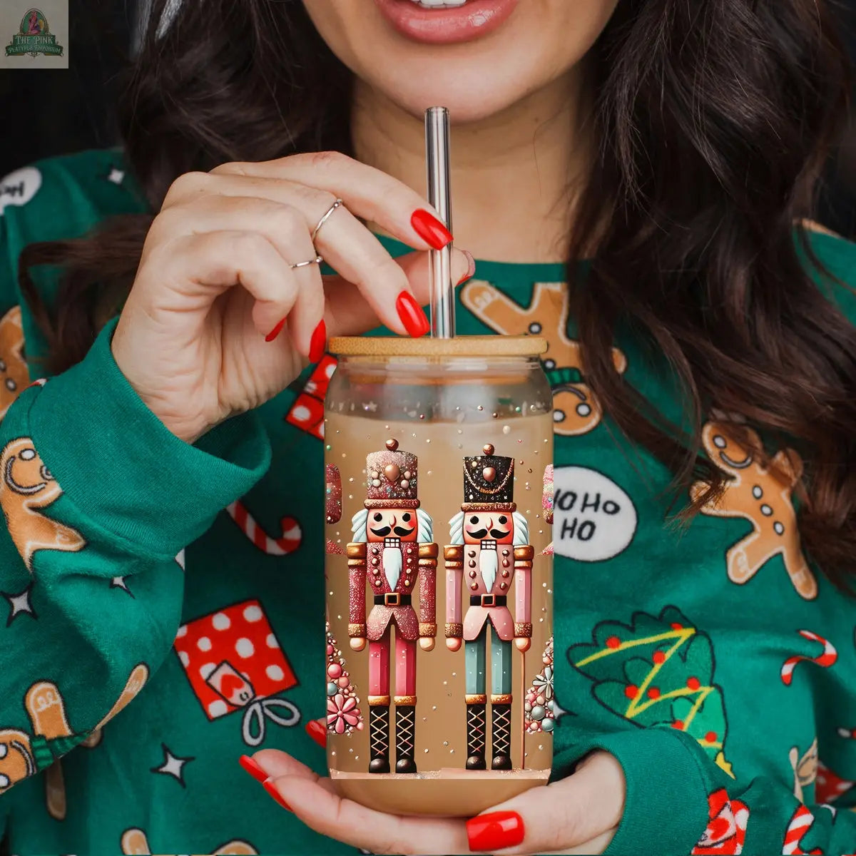 A woman with red nail polish sips iced coffee from the "Nutcracker Glam Soldier" holiday glass tumbler, featuring nutcracker designs. She wears a green sweater adorned with gingerbread men, presents, and Christmas trees.