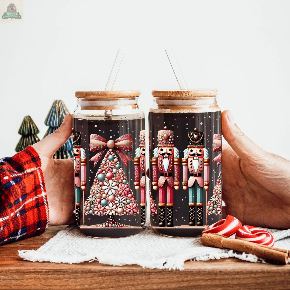 Two hands hold festive Nutcracker Glam Soldier holiday glass jars with wooden lids and straws. The tumblers, featuring Nutcracker designs, rest on a wooden table with a white cloth, candy canes, and small decorative trees in the background.