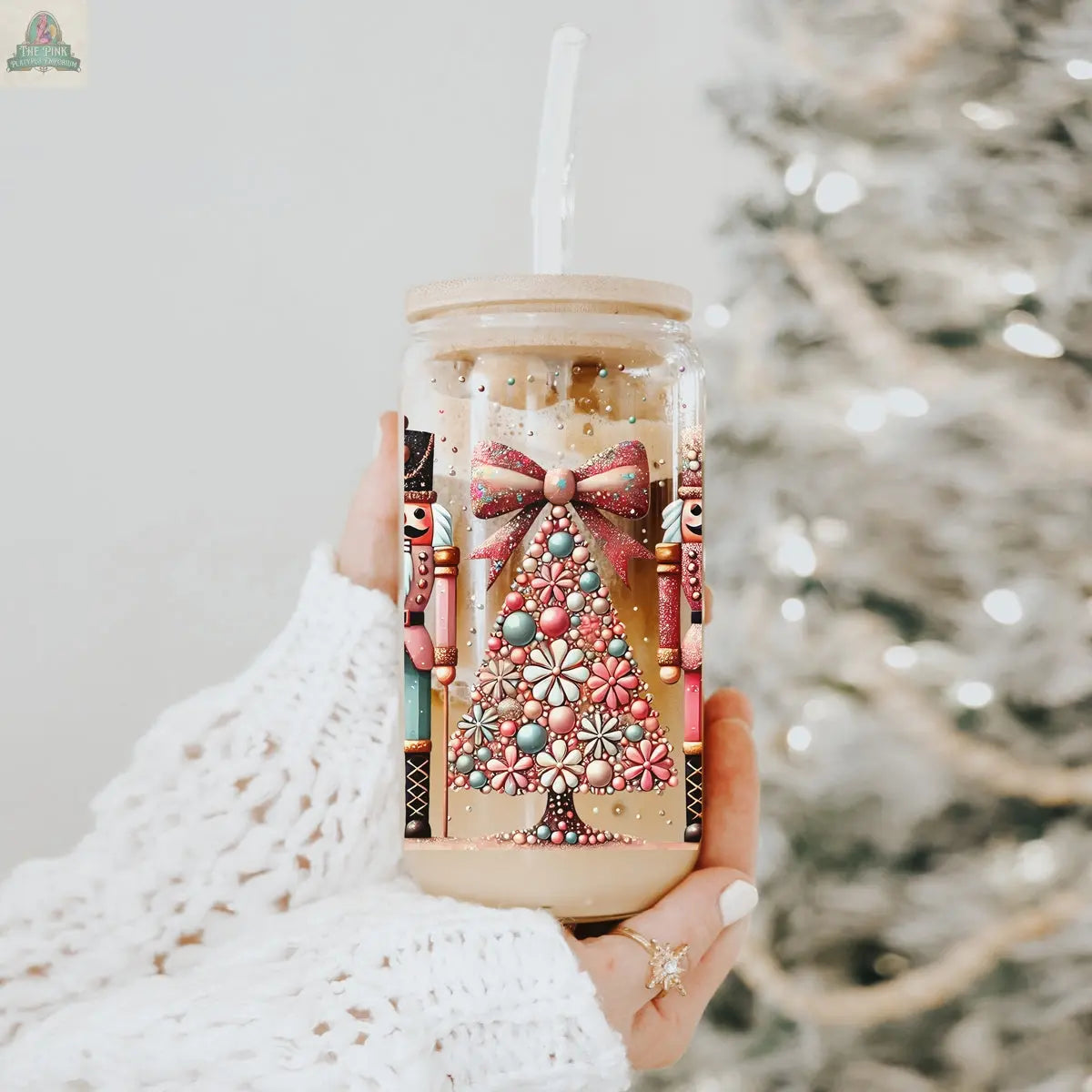 A hand in a cozy white sweater holds the Nutcracker Glam Soldier holiday glass jar filled with iced coffee, featuring a festive Christmas tree, nutcrackers, and a pink bow. A lit Christmas tree is beautifully blurred in the background.