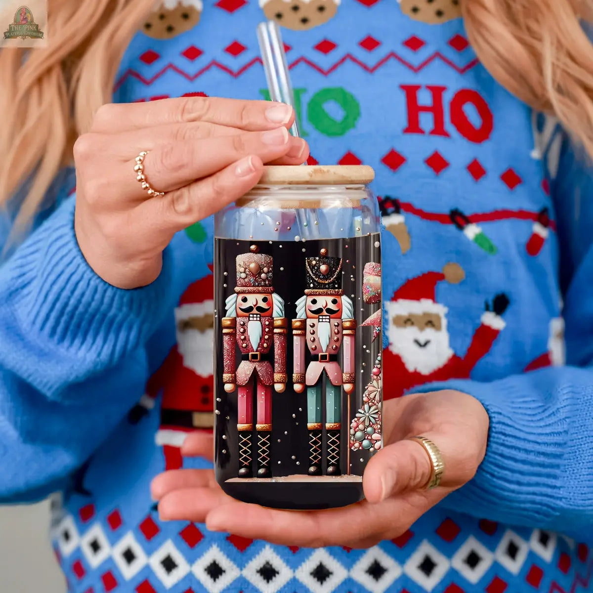 A person wearing a festive blue sweater with Santa faces holds a Nutcracker Glam Soldier glass tumbler with colorful nutcracker designs, a bamboo lid, and a clear straw.
