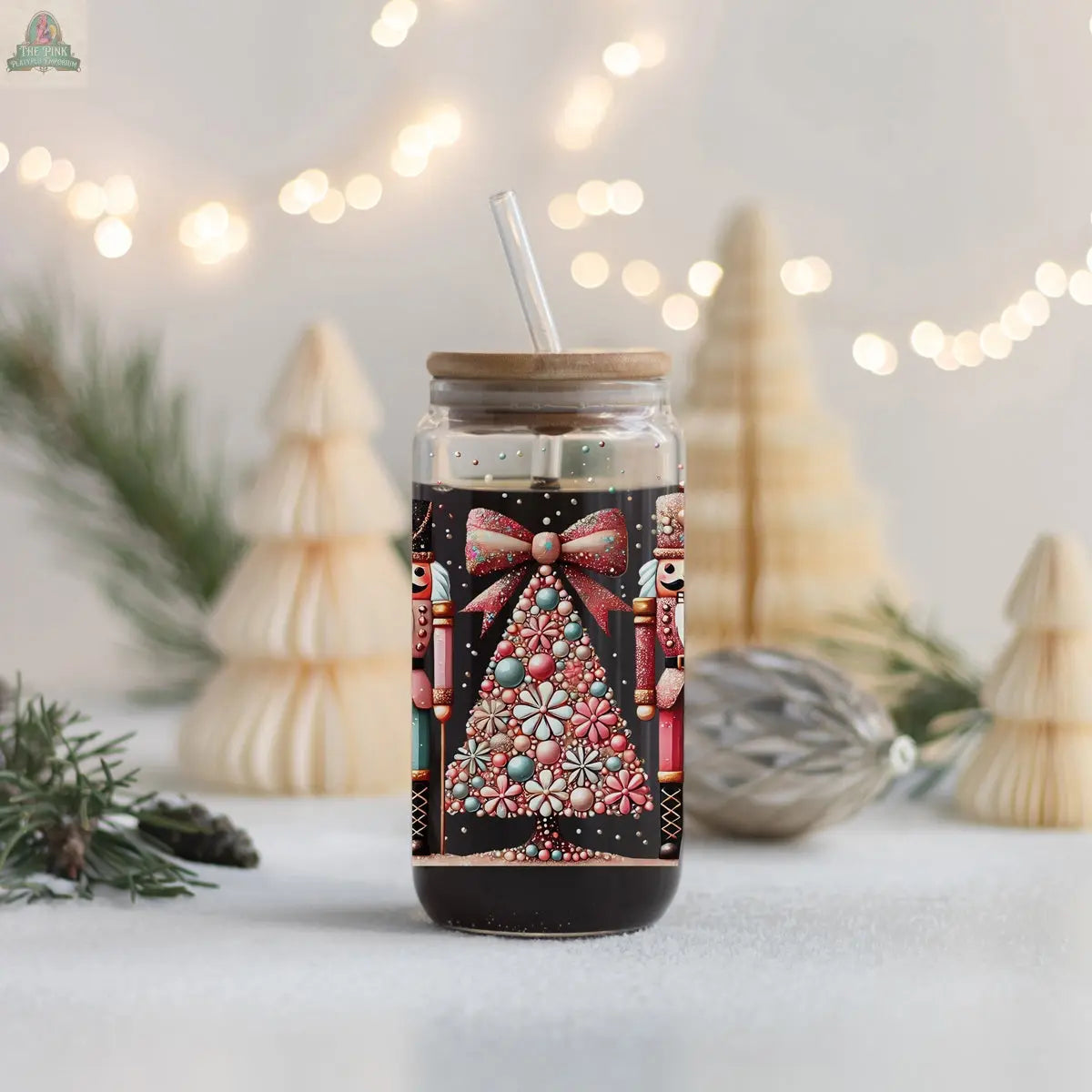 A glass jar with a wooden lid and straw is filled with a dark drink. This Nutcracker Glam Soldier glass tumbler features a festive design, while blurred holiday lights create a cozy holiday vibe in the background.