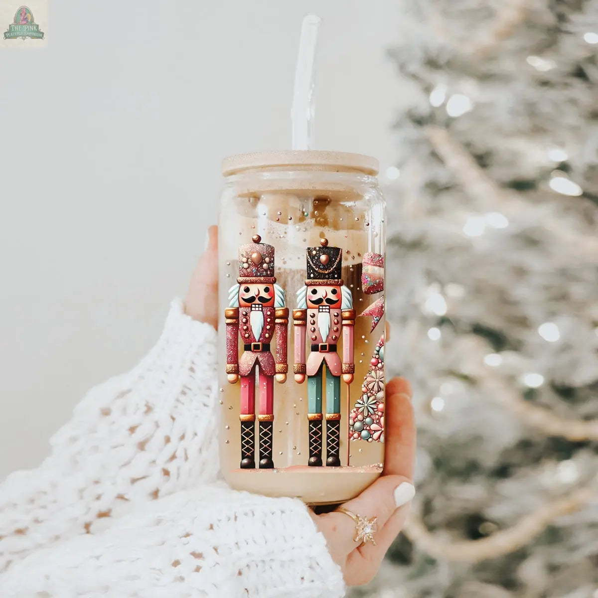 A hand in a white knitted sleeve holds the Nutcracker Glam Soldier tumbler, featuring festive holiday designs, with a blurred, lit Christmas tree in the background.