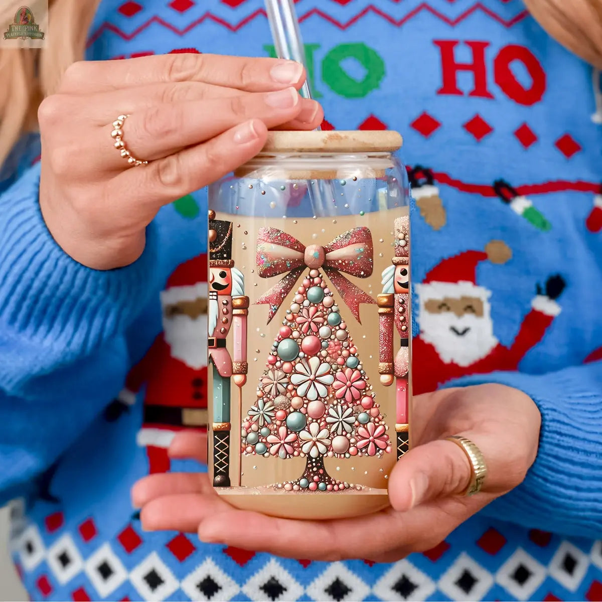 Wearing a festive blue HO HO HO Santa sweater, someone holds an iced coffee in the Nutcracker Glam Soldier glass tumbler, featuring nutcrackers and a jeweled Christmas tree for extra holiday drinkware charm.