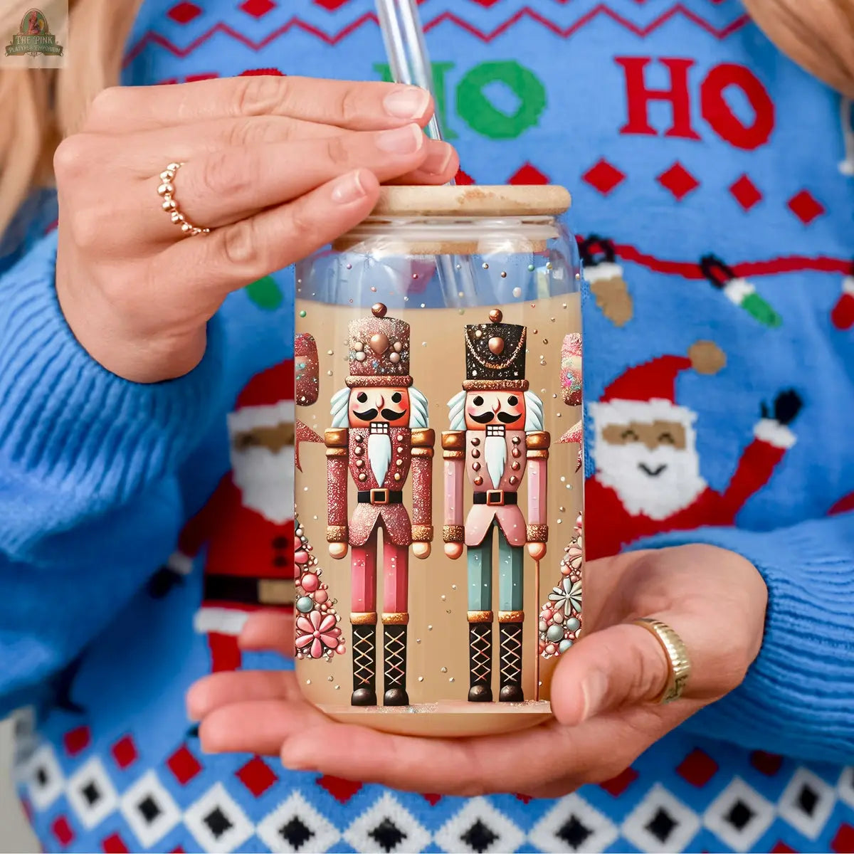 A person in a blue holiday sweater with Santa faces holds the Nutcracker Glam Soldier glass tumbler, decorated with two colorful nutcracker designs. This holiday drinkware includes a wooden lid and clear straw.