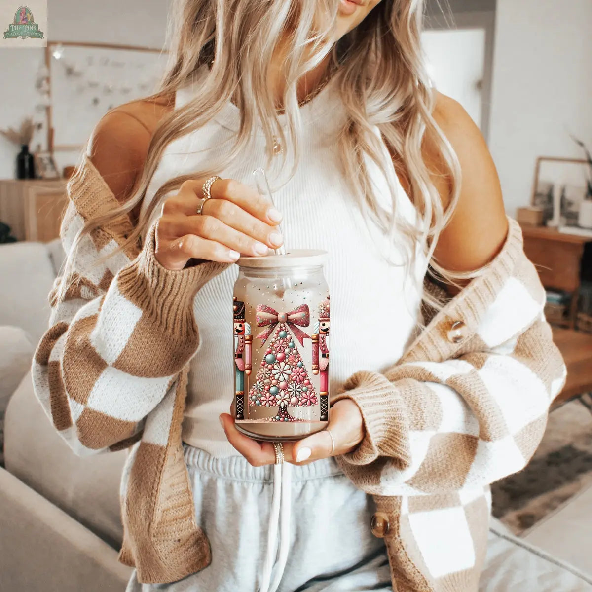 A woman with long, wavy blonde hair holds a Nutcracker Glam Soldier glass tumbler decorated with a pink holiday tree and bow. She wears a white top, tan and white checkered cardigan, and gray drawstring pants in a cozy living room.