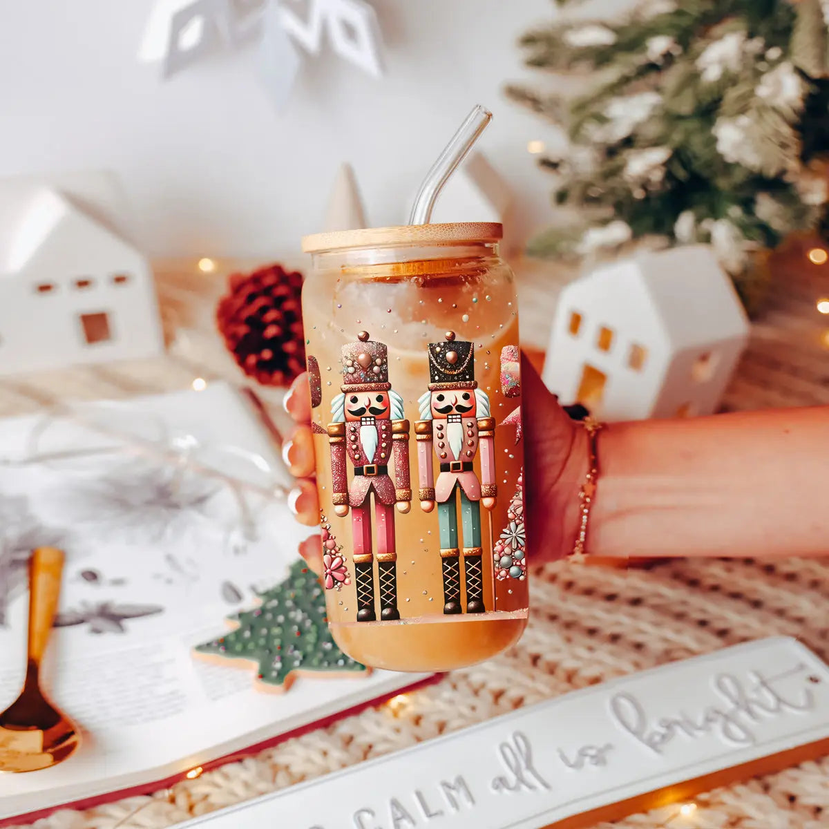A hand holds the Nutcracker Glam Soldier Christmas glass tumbler with bamboo lid and straw, featuring nutcracker designs. In the festive background are an open book, a cookie, and white house ornaments.