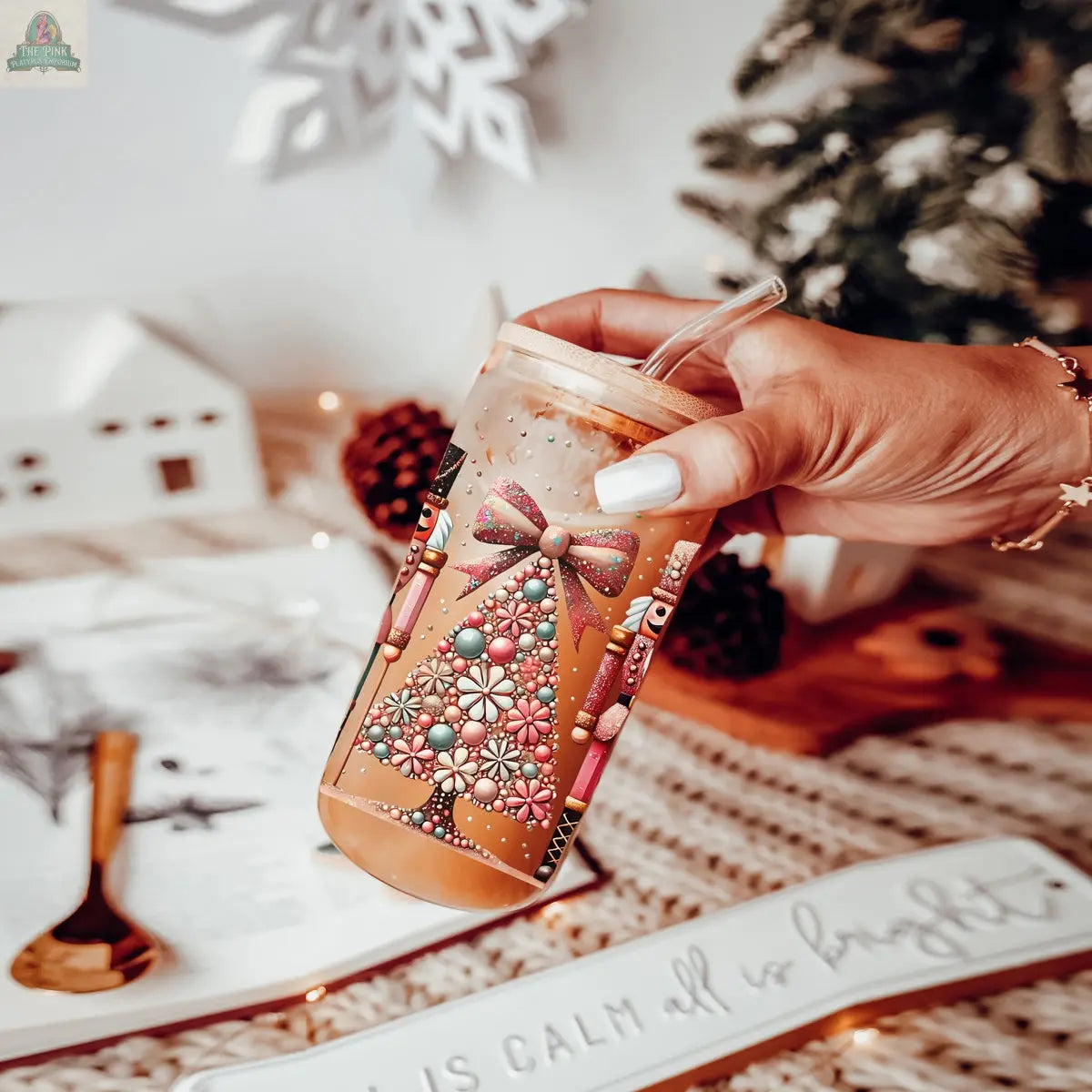 A hand holds an iced coffee in a festive "Nutcracker Glam Soldier" Christmas glass tumbler, surrounded by cozy holiday decor and a plate that reads “All is calm all is bright.”.