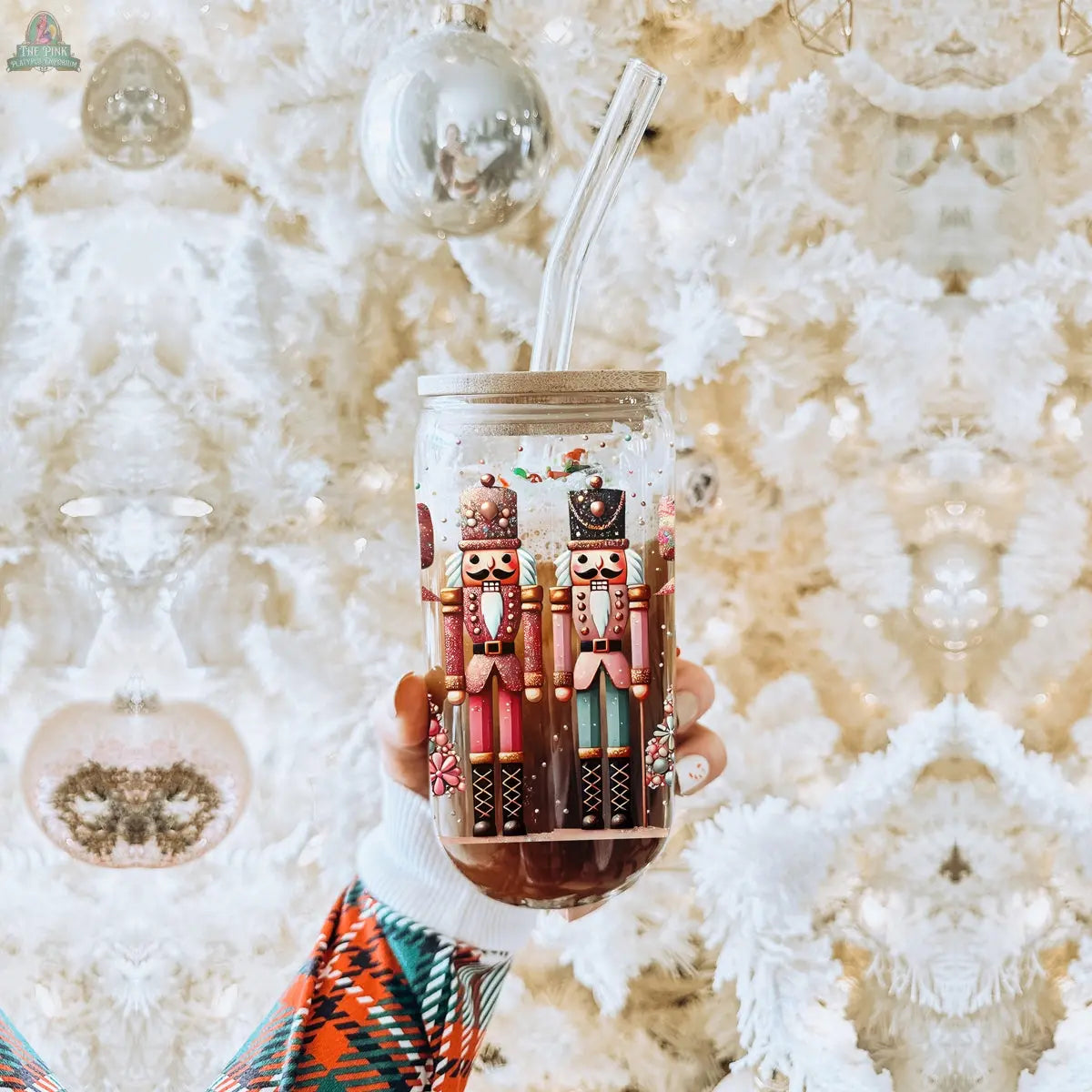 A hand holds the Nutcracker Glam Soldier glass tumbler with a clear straw and festive nutcracker design. A white Christmas tree with ornaments is in the background, highlighting this holiday drinkware.