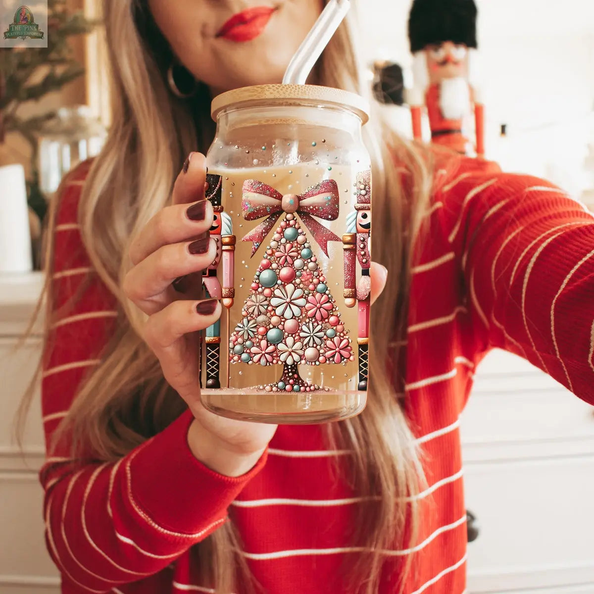 A woman in a red and white striped sweater holds the Nutcracker Glam Soldier tumbler, featuring festive Christmas trees, bows, and nutcracker designs. A nutcracker figurine in the background enhances the holiday charm.