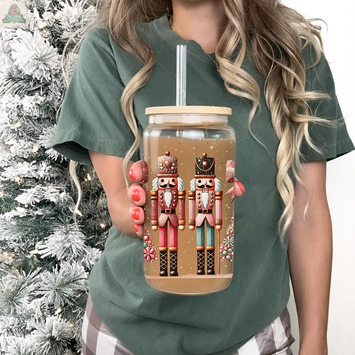 A woman in a green t-shirt holds a Nutcracker Glam Soldier tumbler with a bamboo lid and straw, featuring two vibrant nutcrackers. A snowy artificial Christmas tree creates a festive background.
