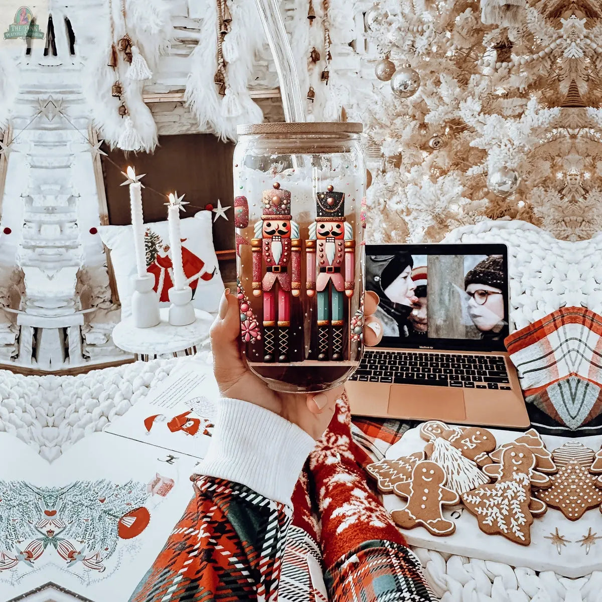 A person in festive pajamas holds a Nutcracker Glam Soldier tumbler featuring Nutcracker figures. Gingerbread cookies, a laptop playing a black-and-white movie, a coloring book, candles, and a white Christmas tree are in the background.