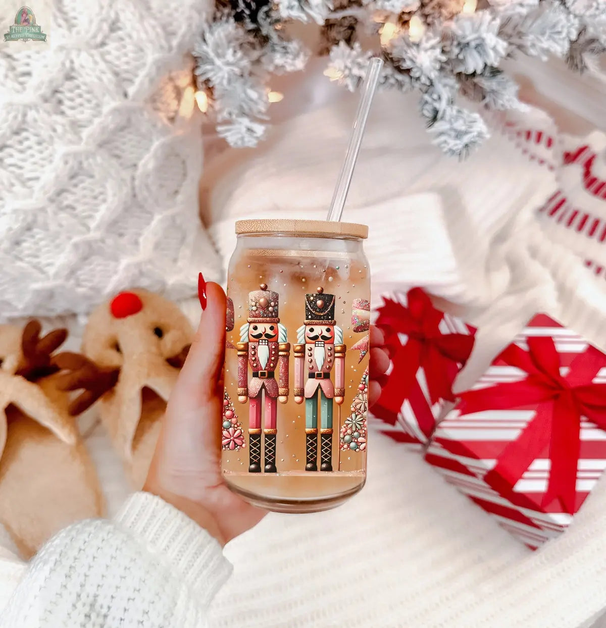 A hand holds a Nutcracker Glam Soldier tumbler with a light brown drink and straw. Fuzzy reindeer slippers, a red-and-white gift, and holiday décor appear in the background—perfect festive drinkware for cozy Christmas moments.