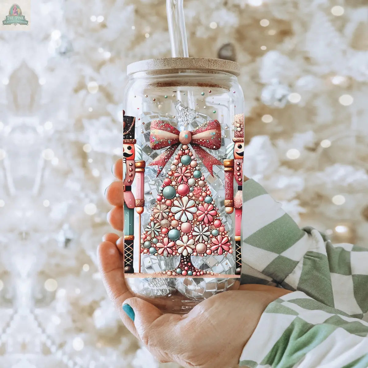 A person holds the Nutcracker Glam Soldier Christmas glass tumbler with a straw, adorned with a jeweled tree, pink bow, and nutcrackers. Festive drinkware sparkles as a blurred white and pastel Christmas tree decorates the background.