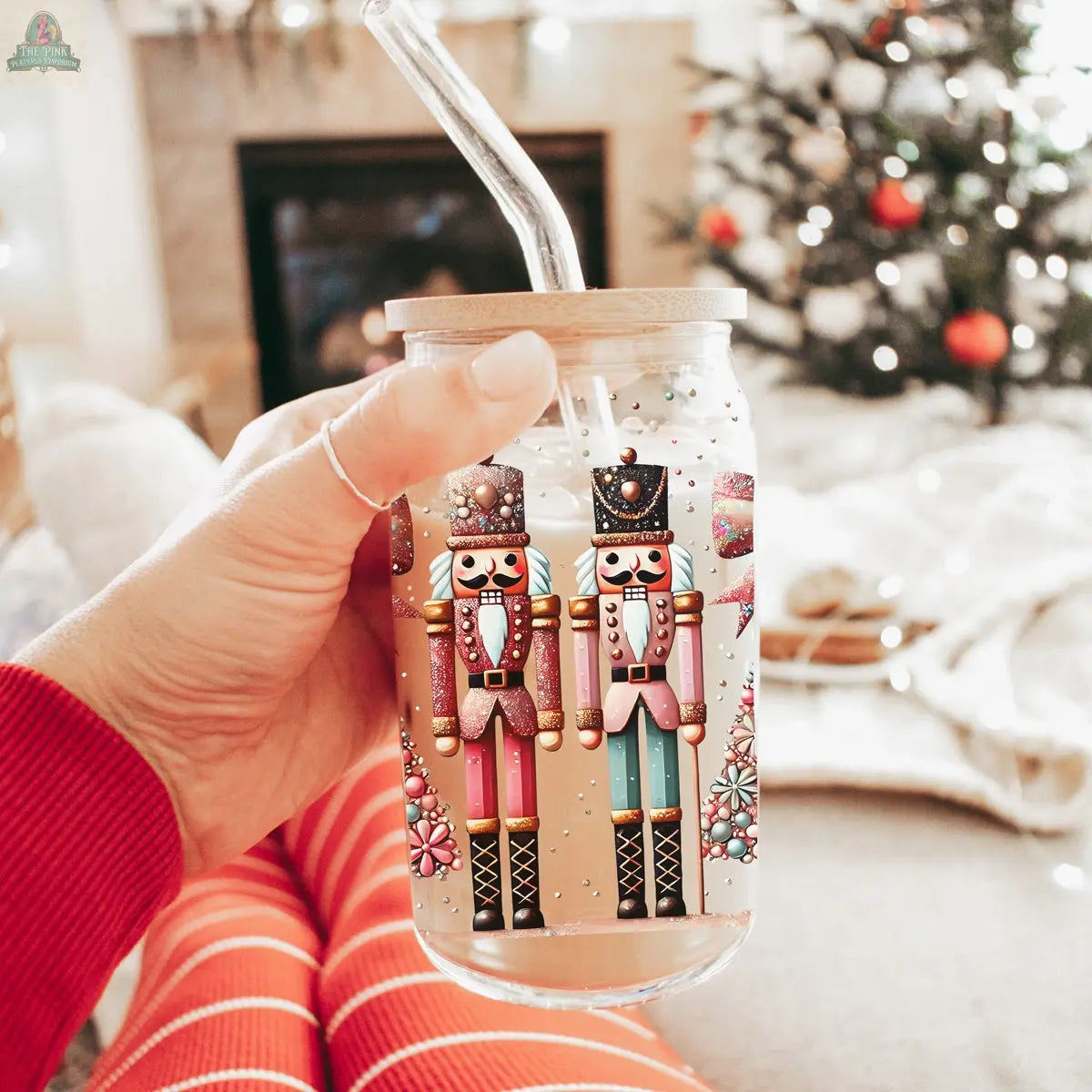 A hand holds the Nutcracker Glam Soldier tumbler with a straw, featuring colorful holiday designs, in front of a cozy living room with a Christmas tree. The person wears red striped pajamas, enhancing the festive drinkware scene.