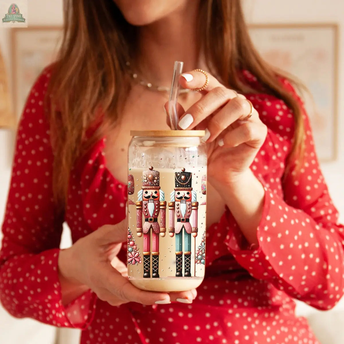 A woman in a red polka-dot dress holds a Nutcracker Glam Soldier tumbler with lid and straw, perfect as festive holiday drinkware. The focus is on the Christmas tumbler, with her face partially out of frame.