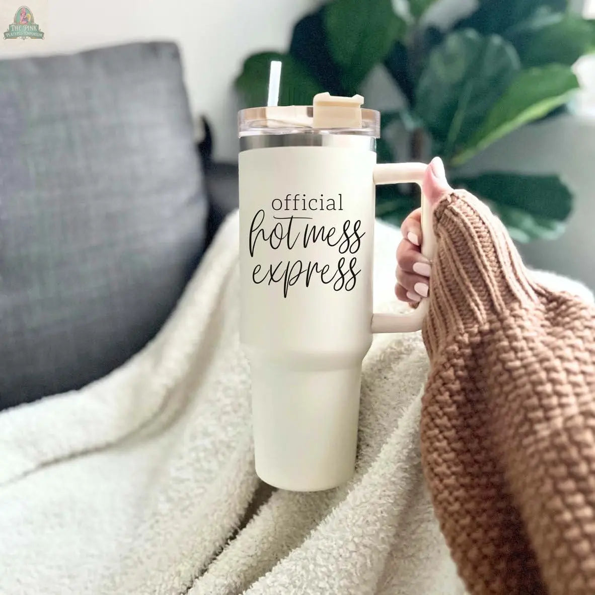 Someone in a brown sweater holds the Official Hot Mess 40oz tumbler with a straw, featuring the text "Official Hot Mess Express." A cozy blanket, sofa, and green plant appear in the background.