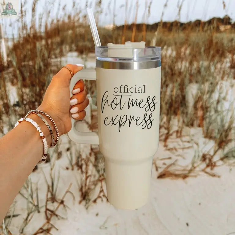 A hand holds the Official Hot Mess 40oz tumbler with a clear lid and straw on a sandy beach with tall grass. This insulated cup keeps drinks cool while showing off your style.