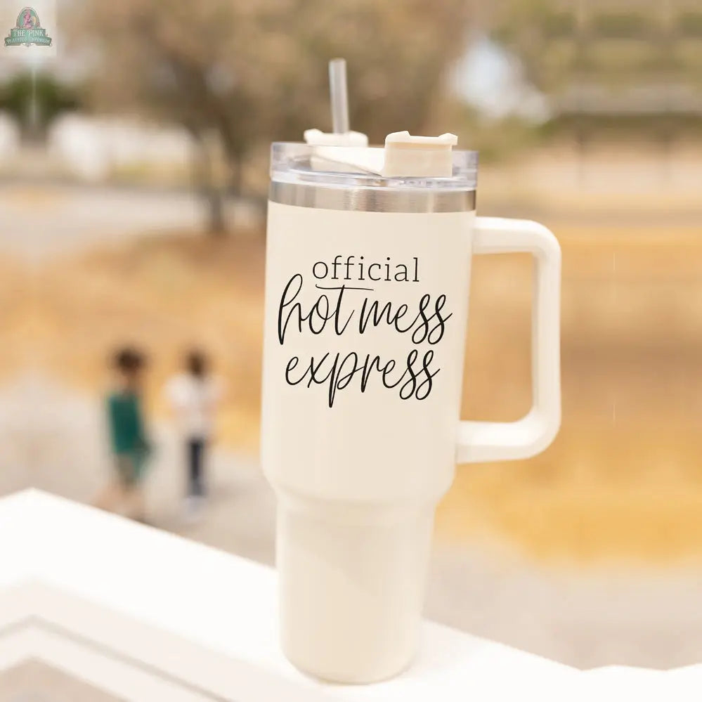 The Official Hot Mess 40oz white insulated cup with a handle and straw sits on a ledge, displaying "Official Hot Mess Express" in black script. The background is blurred, revealing two children and a dry outdoor area.