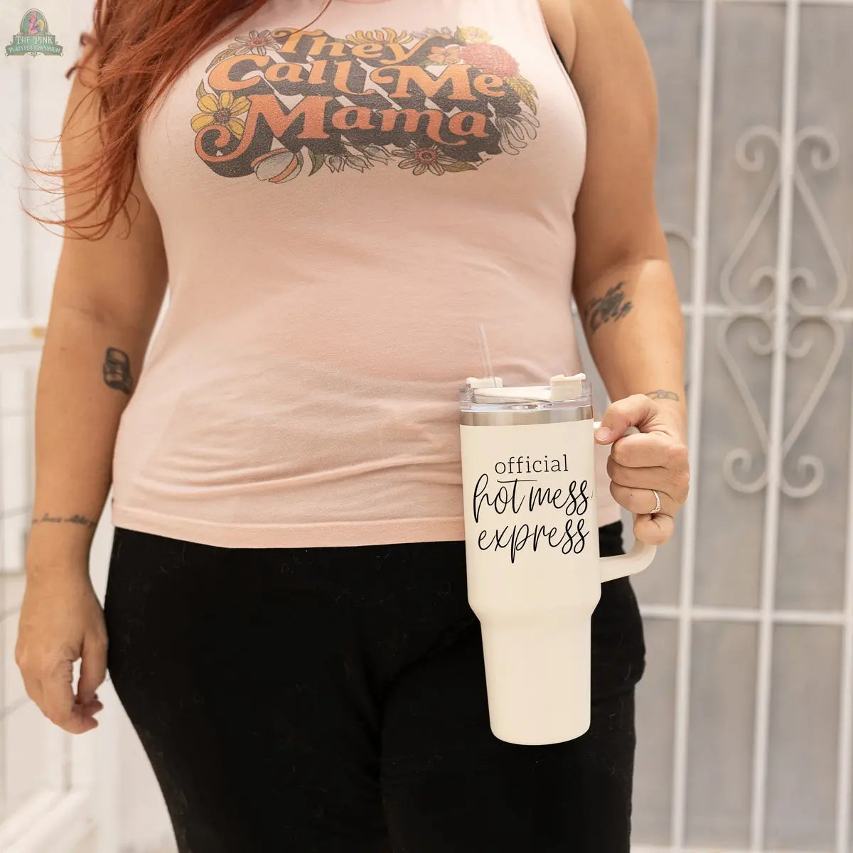 Wearing a “They Call Me Mama” tank, a person holds the Official Hot Mess 40oz tumbler. A white gate appears in the background.