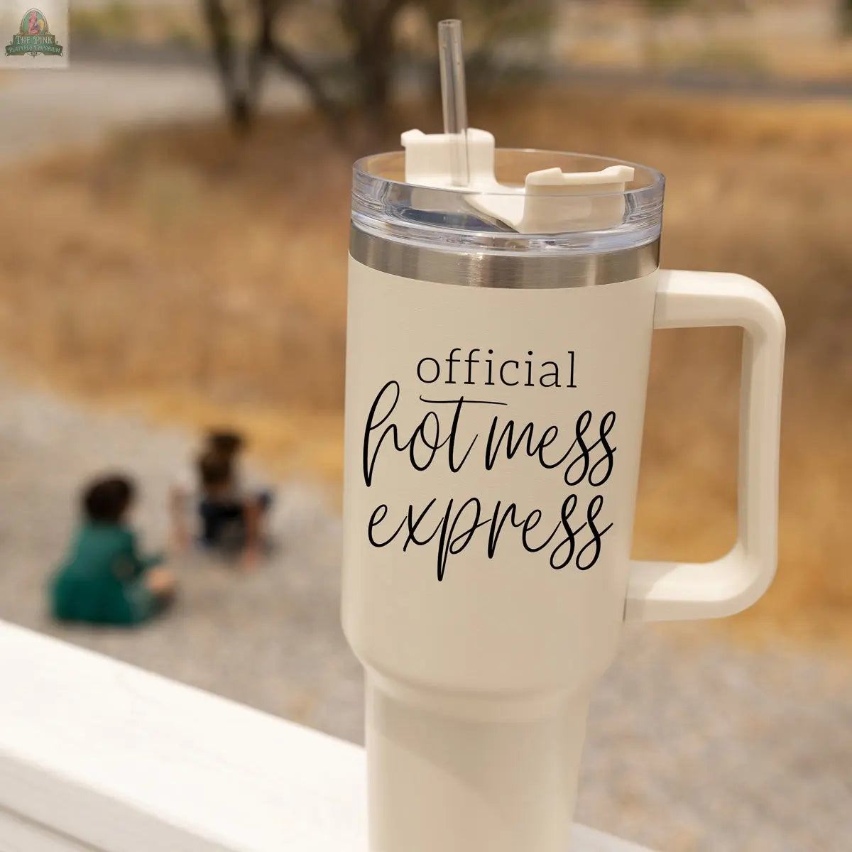 A cream Official Hot Mess 40oz insulated tumbler with a straw and handle rests on a railing. "Official Hot Mess Express" is printed on the cup. Two children are visible, blurred, sitting outside on the ground.