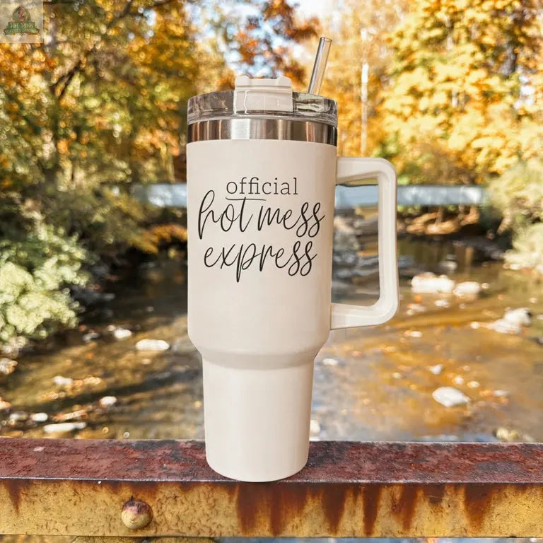 The Official Hot Mess 40oz white tumbler with handle and straw rests on a rustic railing. Its insulated design and “Official Hot Mess Express” text stand out, while a river and autumn trees in the background create a cozy feel.