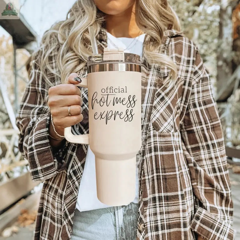 A woman with wavy blonde hair wears a brown plaid shirt and holds the Official Hot Mess 40oz tumbler. She stands outdoors, with trees and a blurred background.