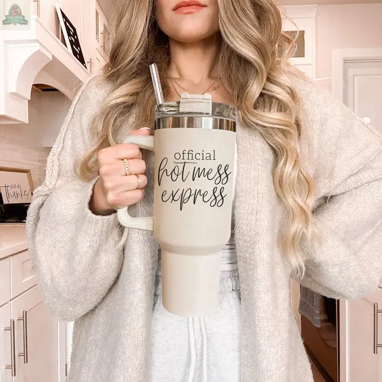 A woman with long wavy blonde hair in a beige cardigan holds the Official Hot Mess 40oz tumbler in a bright, modern kitchen.