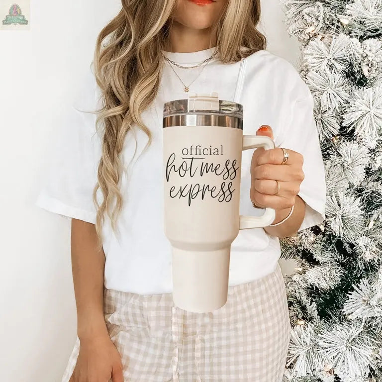 A woman with long wavy hair holds the Official Hot Mess 40oz tumbler, wearing a white shirt and plaid pants beside a frosted Christmas tree.