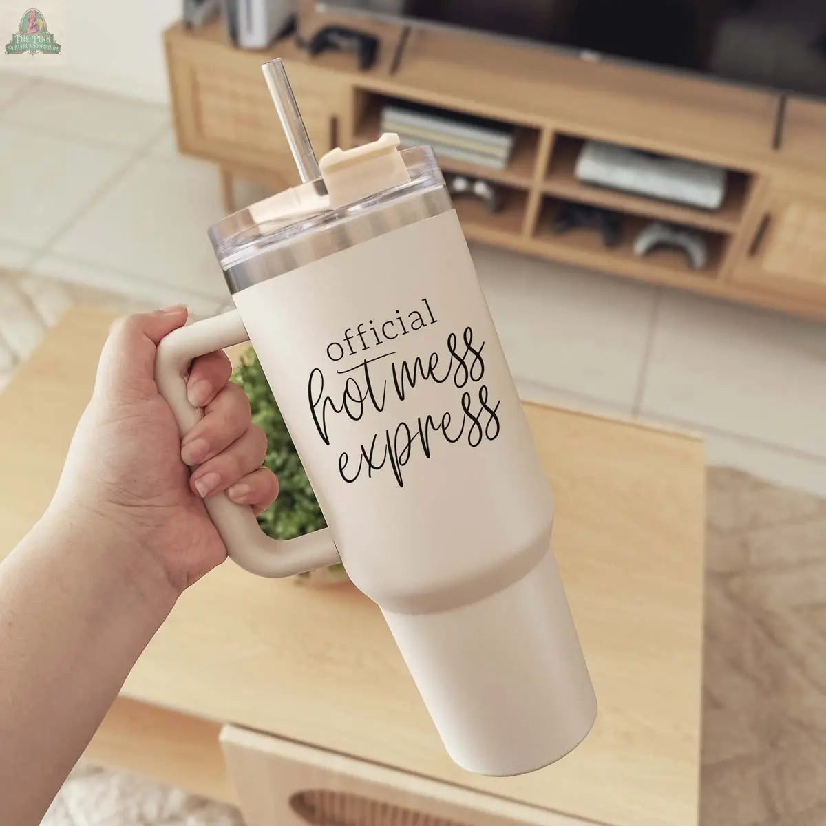 A hand holds the Official Hot Mess 40oz beige tumbler with a lid and straw, featuring "Official Hot Mess Express" in black script. A wooden coffee table and TV stand appear in the background.