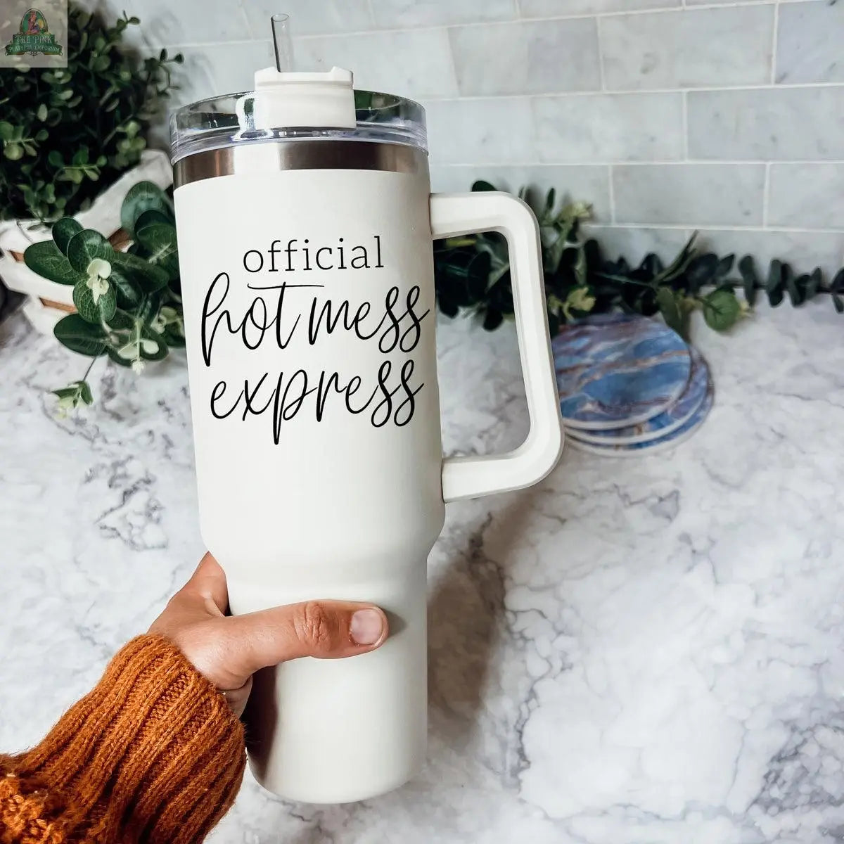 A hand in an orange sweater holds the Official Hot Mess 40oz tumbler, labeled “Official Hot Mess Express” in black script. The insulated cup rests on a marble counter with green plants in the background.