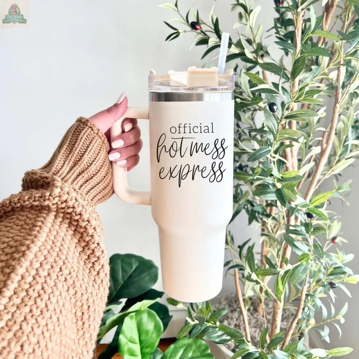 A hand in a brown knit sweater holds the Official Hot Mess 40oz tumbler, with plants visible in the background.